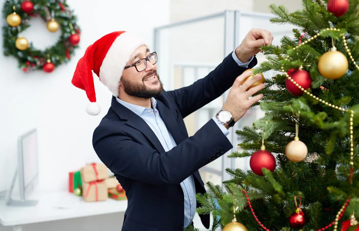 Employé en costume avec un bonnet de Noël décorant un sapin de Noël dans un bureau festif, avec des guirlandes et des décorations en arrière-plan.