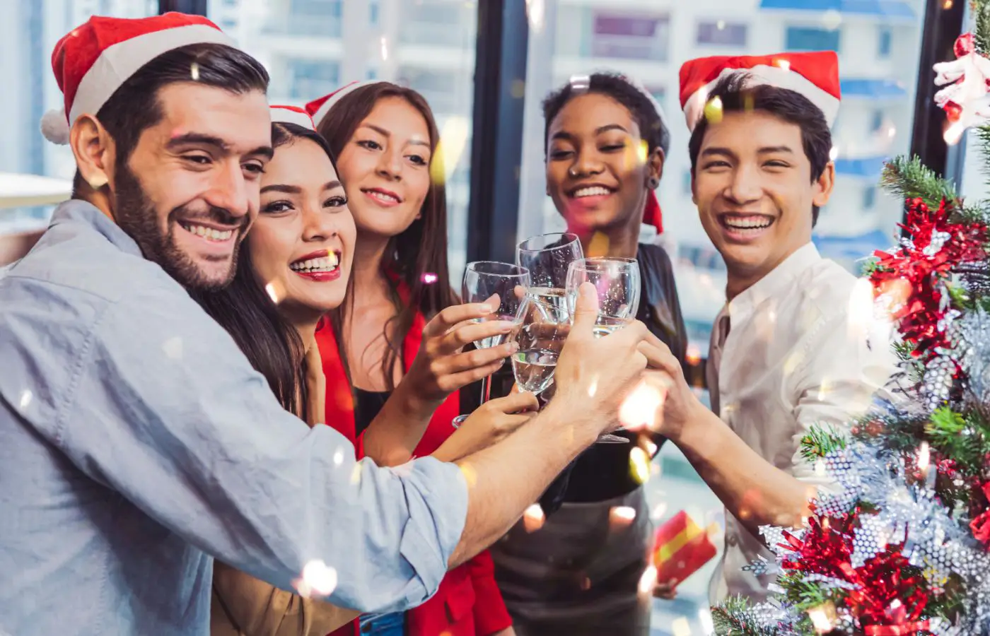 Groupe de collègues souriants célébrant Noël en entreprise, portant des bonnets de Noël et trinquant près d’un sapin décoré.